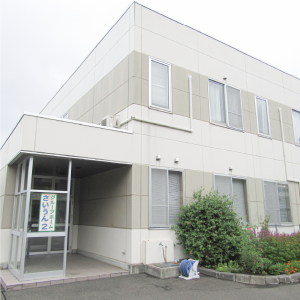 2階建ての介護施設の外観。白系の外壁に複数の窓があり、玄関前に緑の植栽がある。